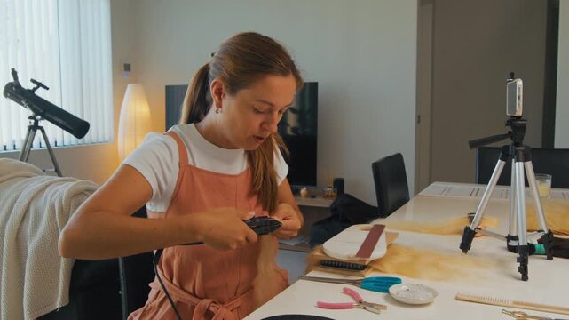 A woman sits at a table with hair styling tools and a camera. She holds a piece of hair while preparing for her recording session.