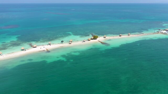 Aerial drone video of a long, narrow white sandbar in the Caribbean Sea, featuring turquoise water, coral reefs, and rustic palapas under a bright tropical sun