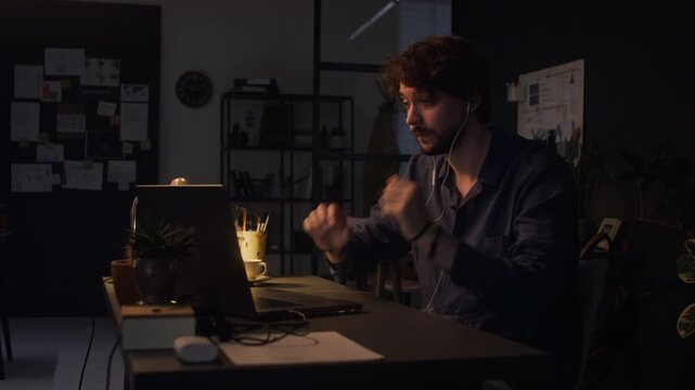 Caucasian young adult male employee putting on earphones and speaking during video call on laptop in dark office at night. Video suits corporate communication and remote work marketing campaigns
