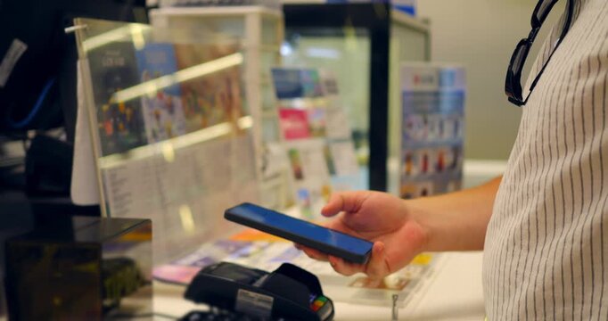 Mobile payment at retail checkout with customer holding smartphone near card terminal and cashier counter, contactless transaction in shop interior with display stand and receipt counter visible.