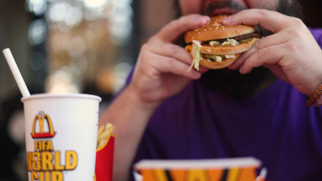 Tbilisi, Georgia - July 4, 2026: Defocused photo. Unknown fat man in purple t-shirt eats Big Mac burger. Cup with McDonald's Golden Arches, logo of FIFA World Cup with sweet drink straw out of focus
