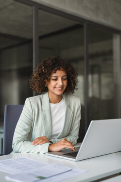 Curly hair latin young business woman working on laptop computer reading financial document report in office. Accountant entrepreneur manager businesswoman doing paperwork analysing using pc. Vertical