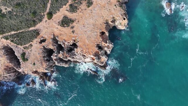 Aerial drone top down shot booming up over the rugged coastal cliffs of Sagres, Portugal, part of the Rota Vicentina's Fisherman's Trail. Turquoise ocean waters crash against the shore.