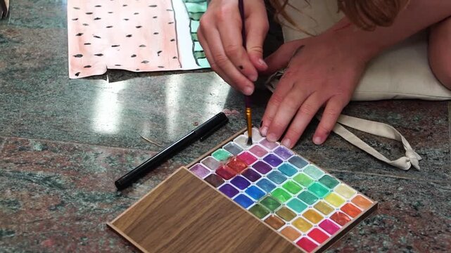 Сhild&rsquo;s hands carefully dipping a brush into a watercolor palette and painting on a sheet of paper with a watermelon pattern. The palette contains many bright colors arranged in a wooden box.