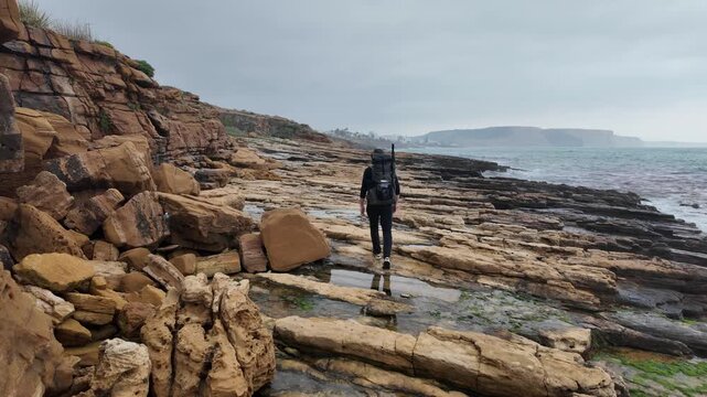 A lone hiker with a backpack traverses a rugged, rocky coastline, part of the scenic Fisherman's Trail. The cloudy sky hangs over the vast ocean as waves crash onto the shore.