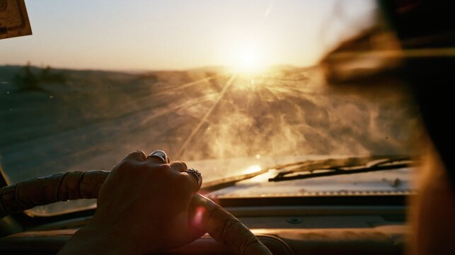 Person with rings on fingers grips the steering wheel while driving through a bright desert landscape during golden hour sunset