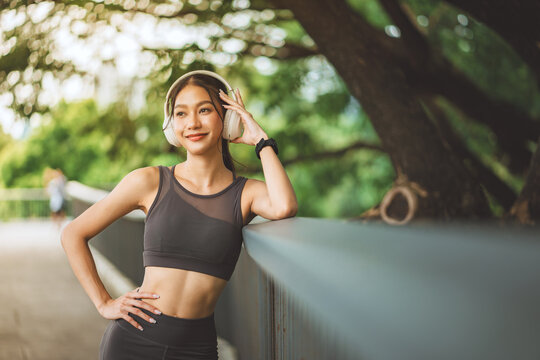Happy Asian woman wearing headphones and sportswear leaning on bridge railing at park, smiling fitness female athlete listening to music during outdoor workout break for active lifestyle, healthy