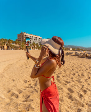 Rear view of a beautiful young Cuban woman in a red bikini and straw hat recording a video blog or making a video call, with the recording image on her smartphone screen on a sunny beach.