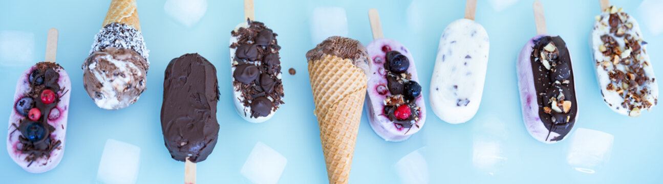 Colorful row of homemade yogurt and milk ice pops. Vanilla and chocolate decorated with nuts and frozen berries with ice cubes on bright blue background. Perfect for a summer children's birthday party