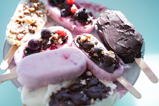 Colorful assortment of homemade yogurt popsicles. Vanilla and chocolate decorated with nuts and frozen berries on ice blue background. Perfect for summer children's birthday party. Overhead close-up s