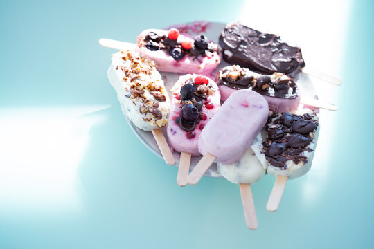Colorful assortment of homemade yogurt popsicles. Vanilla and chocolate decorated with nuts and frozen berries on ice blue background. Perfect for summer children's birthday party. Overhead close-up 