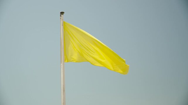 Yellow flag waves on flagpole against blue sky in sunny weather. Flag displayed by lifeguards on developed beach restricting swimming in sea and ocean. Deteriorating weather conditions for recreation.