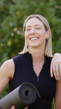 Vertical video: Smiling woman in black top holding rolled mat, reacting to pal on shoulder in park