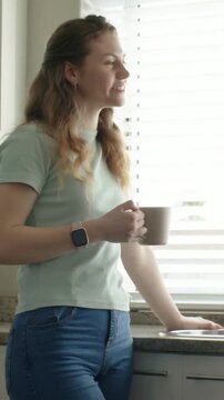 Vertical video: Reacting to blinds, woman in green tee holding mug tapping smartwatch in kitchen