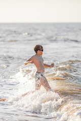 Fototapeta premium Blond boy in black swim goggles runs through foamy sea surf as waves splash around him on sunny beach.
