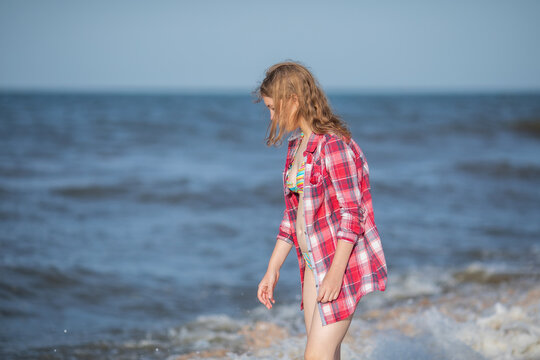 Girl in red plaid shirt standing by sea waves on beach.