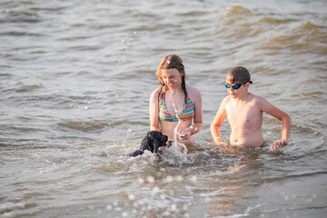 Fototapeta premium Teenage girl in striped bikini and blond boy in swim goggles play with black spaniel in sea water.