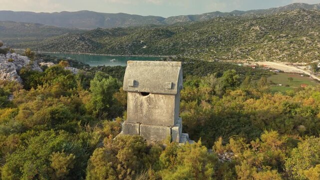 Ancient Lycian stone tomb stands on green rocky hill overlooking Kekova bay, Turkey. 4K drone view captures historic Mediterranean sarcophagus surrounded by lush bushes in Kekova