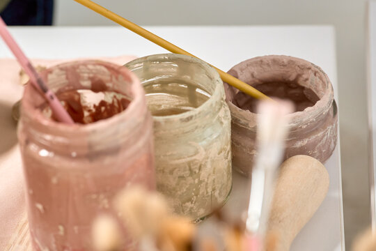 Close-up glass jars filled with colorful clay slip or engobes for pottery decoration. Authentic artist studio workspace with brushes and creative supplies.