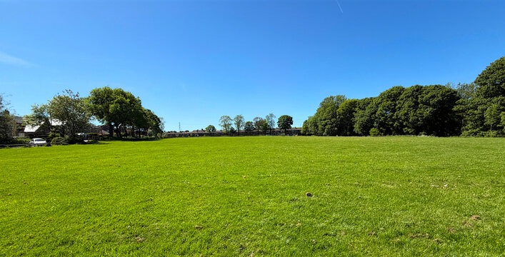 A wide, open grassy field spreads out beneath a clear blue sky, with trees lining the edges and casting soft shadows across the vibrant green grass in Shelf Hall Park, Shelf, Bradford, UK.