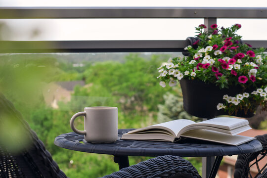 Eye-level shot showing white mug and open book on black metal table with hanging flower basket and blurred green tree background
