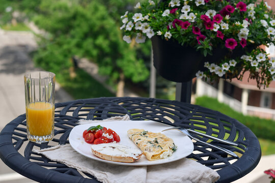 High angle close-up showing breakfast plate with omelet, bagel, tomato salad and glass of orange juice on black metal table with hanging flower basket
