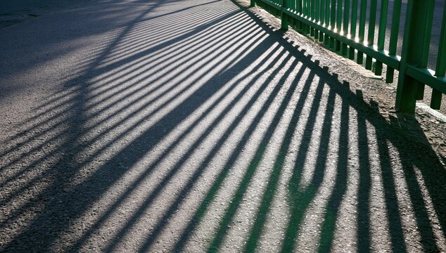 Green fence casting long shadows on road in sunny outdoors