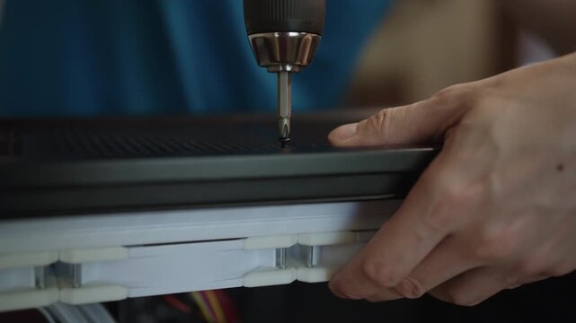Close-up view of skilled worker hands using cordless electric screwdriver to fasten into component during assembly process of complex electronic device in factory. Shooting in slow motion
