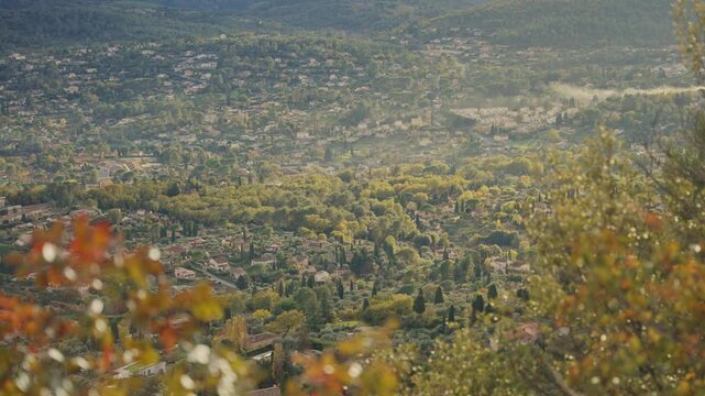 Autumn leaves frame a sprawling small village or town in France's Provence. Perfume country during beautiful peaceful day with green trees and quintessential view on medieval villages in France