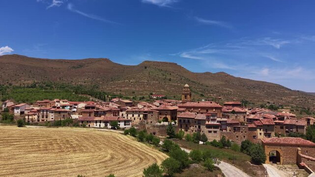 Aerial view of Mirambel medieval stone village with terracotta rooftops and church tower, bordered by harvested wheat field and mountain landscape, Teruel, Aragon, Spain