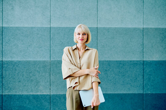 Young blonde female professional holding digital tablet, smiling, standing confidently in modern office building workplace