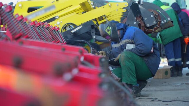 Agricultural mechanic in a welding mask repairing a large red and yellow seeder machine outdoors in winter