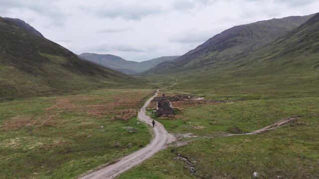 Aerial drone push forward shot of a hiker walking the West Highland Way in Scotland. He walks a long winding road into a valley surrounded by high mountains, crossing the Tigh-na-sleubhaich ruin.