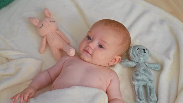 Baby girl lying on soft blanket with knitted bunny and bear, bare chest visible, smiling and reaching toward toy in studio; calm.
