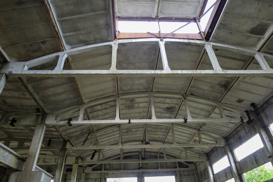 The massive reinforced concrete ceiling of an old abandoned industrial hangar or factory workshop shows visible signs of decay. Daylight penetrates inside through an open roof hatch and empty window o