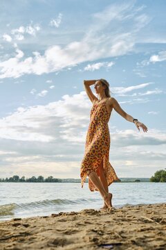 A woman stands barefoot on sandy shores, wearing a bright dress with a bold floral pattern. She playfully raises her arm against the sky with fluffy clouds during a warm afternoon
