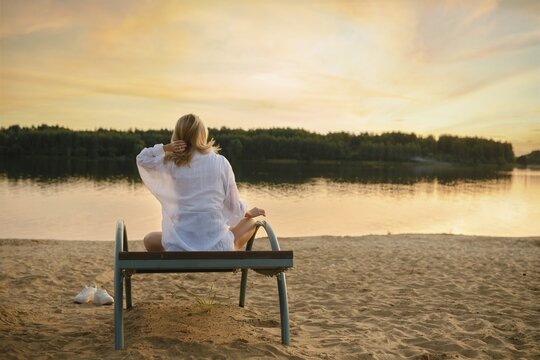A woman sits peacefully on a bench near the riverbank, enjoying the serene sunset. The soft golden light reflects on the water, creating a calming atmosphere perfect for relaxation