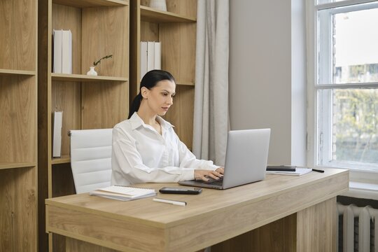 A woman in a white shirt is focused on her laptop in a bright and modern office. The room features wooden shelves filled with books, creating a cozy and productive atmosphere