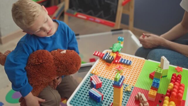 Boy holds a large teddy while placing colorful plastic blocks with his hand on a play table in building; curiosity learning bonding.