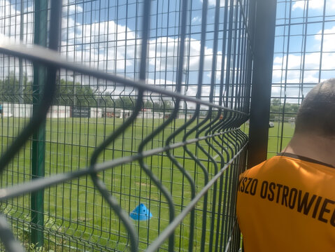 Polska Rączna, 23 maja 2026, 13:50. Widok na mecz piłki nożnej z sektora kibic&oacute;w gości &ndash; drużyny KSZO Ostrowiec. Kibic w pomarańczowej koszulce KSZO  stoi przy drucianym ogrodzeniu i obserwuje mecz.