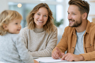 Obraz premium Young family sits together at table. Mom smiles warmly at child. Dad laughs, leaning slightly forward. Child looks up curiously. Scene feels cozy, intimate, and joyful. Perfect for family, parenting