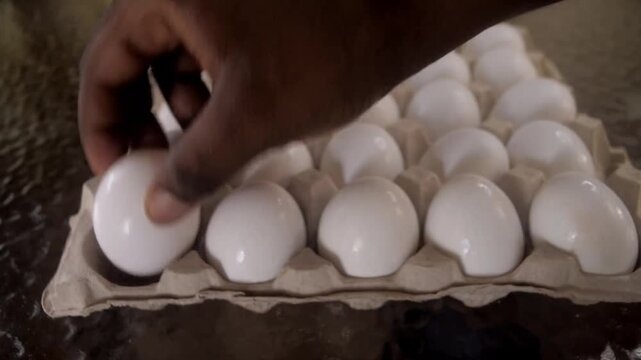 Uses both hands to set down a large and flimsy beige carton of eggs on a clear table to pick out a couple of eggs to cook for an early breakfast.