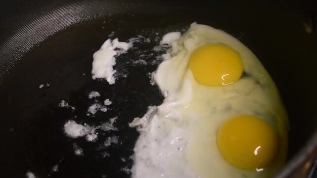 Two raw eggs, freshly cracked into a large black skillet cooking slowly. The oil bubbles and pops underneath the two white eggs. The yolks are whole.