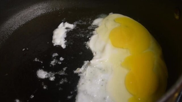 Two raw eggs, freshly cracked into a large black skillet cooking slowly. The oil bubbles and pops underneath the two white eggs. The yolks have broken