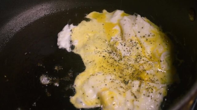 Two raw eggs, freshly cracked into a large black skillet cooking slowly. The eggs are being seasoned with pepper and oregano after cooking for a while