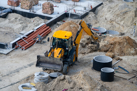 A yellow backhoe loader sits in an active construction site, surrounded by excavated dirt, concrete manhole rings, and stacks of brown pipes.