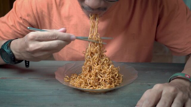 Man eating instant fried noodles with chopsticks at home, close-up comfort food concept, hungry expression, casual lifestyle.