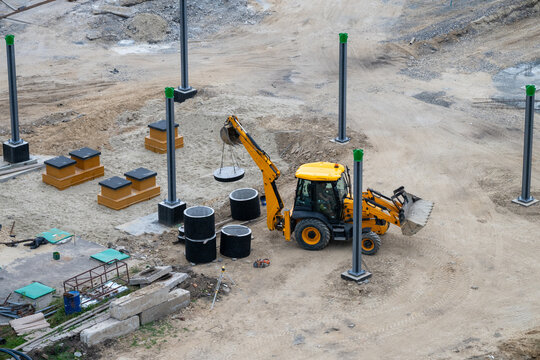a yellow backhoe loader using its rear arm to carefully lift and position a concrete manhole cover among scattered building materials and installed metal pillars.