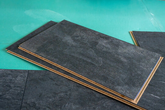 Several dark grey stone-effect laminate flooring planks are stacked on top of each other against a green underlayment, ready for installation.