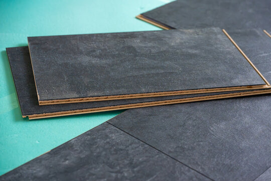 Several dark grey stone-effect laminate flooring planks are stacked on top of each other against a green underlayment, ready for installation.